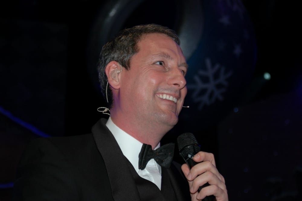 Gert Verhulst, in evening suit, smiling with microphone in hand, embodies the joyful spirit of Studio 100.