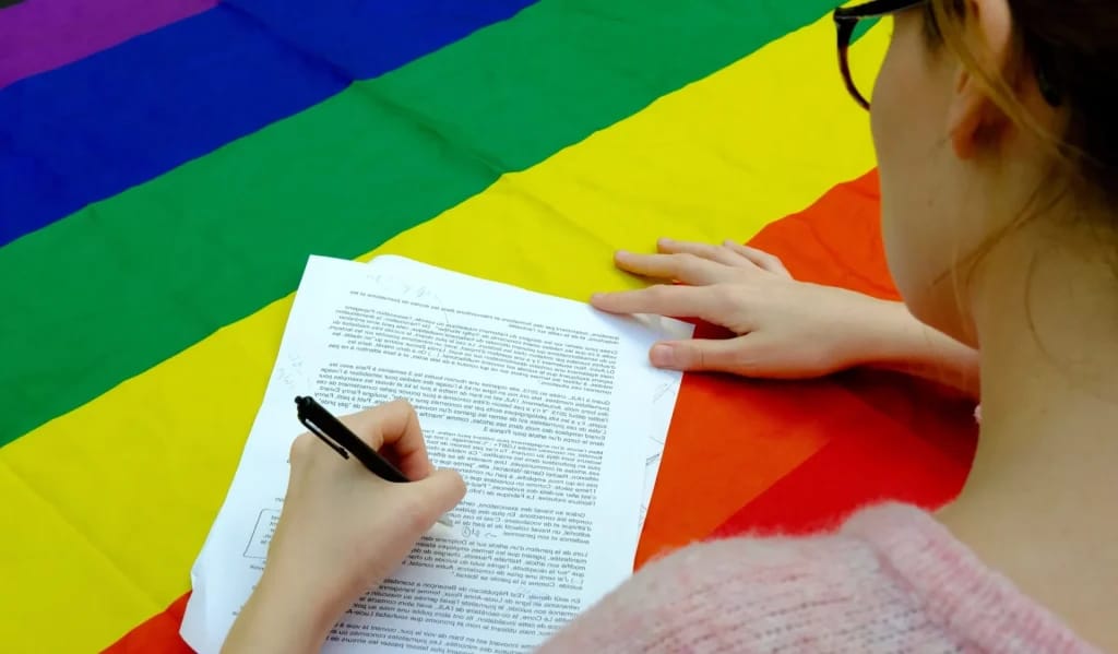 une personne signant un document sur une table couverte par un drapeau arc-en-ciel - Photo d'illustration de l'article Fortune Rachel Garrat-Valcarcel